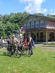 Bild - Zwei Männer in historischen Kleidungen stehen mit Fahrrädern auf einer Wiese vor einem großen, rustikalen Gebäude. Im Hintergrund sind Tische mit Menschen, die im Freien sitzen. Die Atmosphäre ist lebhaft und einladend an einem sonnigen Tag.