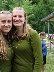 Bild - Zwei junge Frauen stehen nah beieinander, lächeln und tragen grüne Kleidung mit Dekorationen. Im Hintergrund sind weitere Personen und Bäume sichtbar, die eine freundliche, gesellige Atmosphäre vermitteln. Es scheint sich um eine Outdoor-Veranstaltung zu handeln.