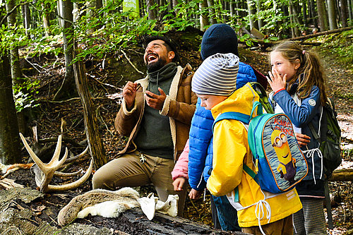 Eine Gruppe von Kindern hört begeistert einem Mann im Wald zu, der fröhlich erzählt. Neben ihnen liegen Tierhörner und ein Stück Tierfell auf dem Boden. Die Kinder sind in Outdoor-Kleidung und zeigen Interesse an der Natur.
