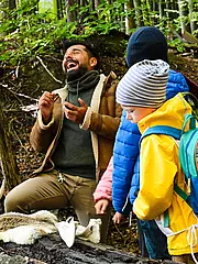 Bild - Eine Gruppe von Kindern hört begeistert einem Mann im Wald zu, der fröhlich erzählt. Neben ihnen liegen Tierhörner und ein Stück Tierfell auf dem Boden. Die Kinder sind in Outdoor-Kleidung und zeigen Interesse an der Natur.