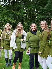 Bild - Eine Gruppe von acht Frauen steht im Freien vor einer grünen Kulisse aus Bäumen. Sie tragen grüne Kleider und haben elfengleiche Ohren sowie dekorative Elemente im Haar. Einige halten Instrumente oder Accessoires. Alle lächeln fröhlich in die Kamera.