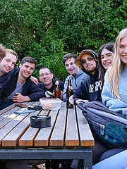 Bild - Eine Gruppe von zehn jungen Menschen sitzt an einem Holztisch im Freien. Sie lächeln und stehen eng zusammen, einige haben   Getränke vor sich. Die Umgebung ist grün mit Bäumen im Hintergrund.
