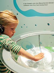 Ein kleines Mädchen mit Kopfhörern untersucht neugierig eine Wasserinstallation. Sie hält ihre Hand über das Wasser, das sanft in einer Schüssel plätschert. Im Hintergrund sind bunte Wände mit Wasser-Themen.