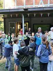 Bild - Eine Gruppe von Kindern steht vor einem Gebäude und hält stolz Medaillen und Zertifikate in der Hand. Sie lächeln und feiern, während einige Erwachsene im Hintergrund beobachten. Das Bild vermittelt Freude und Gemeinschaftsgefühl.