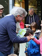 Bild - Eine feierliche Übergabe von Urkunden an Kinder findet statt. Ein Mann überreicht einer lächelnden Mädchen eine Urkunde. Im Hintergrund sind weitere Kinder und Erwachsene zu sehen, die das Ereignis verfolgen. Die Atmosphäre ist fröhlich und festlich.