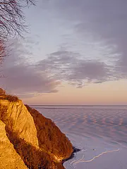 Bild - Ein winterlicher Hang mit Bäumen, der steil zu einer gefrorenen Wasseroberfläche führt. Der Himmel zeigt sanfte Farben des Sonnenuntergangs, während Strukturmuster im Schnee sichtbar sind.