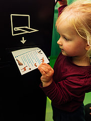 Ein kleines Mädchen steckt einen Zettel mit Bildern von Tieren in einen schwarzen Automaten. Die Umgebung ist grün gestaltet. Das Mädchen trägt ein rotes Oberteil und hat blonde Haare. Es scheint neugierig und konzentriert zu sein.