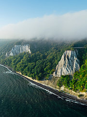 Eine Luftaufnahme zeigt eine Küstenlinie mit beeindruckenden, weißen Klippen, die von üppigem, grünem Wald umgeben sind. Sanfte Wellen treffen auf den Strand, während der Himmel teilweise bewölkt ist. Das Bild vermittelt eine ruhige, natürliche Landschaft.