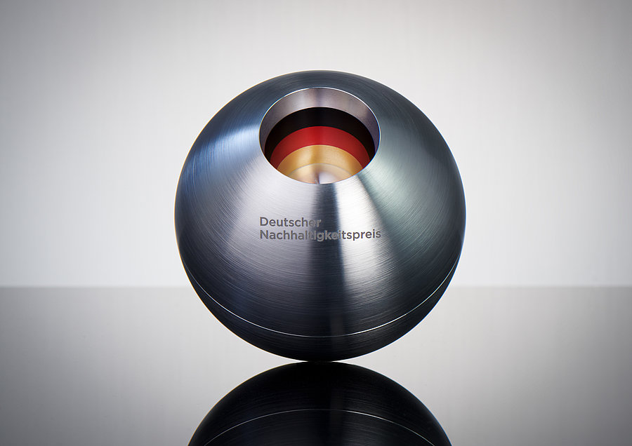 Ein glänzender, runder Metallpokal mit einer zentralen Öffnung, die in verschiedenen Farben gestaltet ist. Der Pokal trägt die Inschrift „Deutscher Nachhaltigkeitspreis“ und steht auf einer reflektierenden Oberfläche.