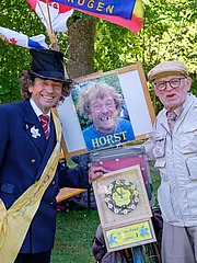 Bild - Zwei Männer stehen im Freien, umgeben von Bäumen. Der erste trägt einen Anzug und einen Hut, der zweite einen Beigen Mantel und eine Schirmmütze. Sie halten ein Schild mit einem Foto einer Person namens Horst und eine Ehrentafel, die einem Wettbewerb gewidmet ist.