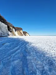Bild - Eine eisbedeckte Küstenlandschaft mit beeindruckenden Klippen im Hintergrund. Der klare Himmel reflektiert das Licht und betont die kalte, unberührte Fläche aus Schnee und Eis. Die Szenerie strahlt Ruhe und Weite aus.