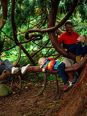 Vier Personen sitzen und spielen in einem Wald. Eine Frau lehnt entspannt an einem Baum, während zwei Kinder mit einem kleinen Jungen auf einem Baumast spielen. Der Junge trägt einen bunten Pullover und steckt an den ausladenden Ästen der Bäume fest.