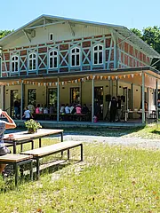 Bild - Ein historisches Gebäude mit einer Veranda, auf der eine Gruppe Menschen sitzt. Im Hintergrund sind Besucher an Tischen zu sehen. Die Umgebung ist grün mit Bäumen und einem klaren Himmel. Fähnchen dekorieren die Veranda, die eine freundliche Atmosphäre schafft.
