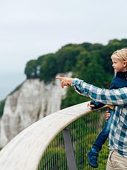 Ein Vater hält sein Kind auf dem Arm und deutet auf die Küste mit weißen Klippen. Im Hintergrund sind weitere Kinder und die grüne Landschaft zu sehen. Die Szenerie vermittelt einen Eindruck von Familientreffen in der Natur.