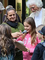 Bild - Eine Gruppe von Kindern versammelt sich um ein Mädchen, das ein Mikrofon hält und auf ein Stück Papier schaut. Sie sprechen miteinander und es scheint, als würden sie etwas Wichtiges oder Lustiges teilen. Eine Frau und ein älterer Mann beobachten sie aufmerksam.
