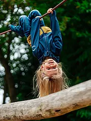 Bild - Ein kleines Kind mit langen Haaren hängt kopfüber an einem Seil in einem Spielplatz. Es trägt einen blauen und gelben Anzug und lächelt fröhlich. Um das Kind herum sind Bäume und eine natürliche Umgebung sichtbar.