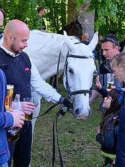 Bild - Auf dem Bild stehen mehrere Erwachsene und Kinder in einem Grünen Park, die einem weißen Pferd nahe sind. Die Kinder streicheln das Pferd, während einige Erwachsene Getränke in der Hand halten. Im Hintergrund sind weitere Pferde und Menschen zu sehen.
