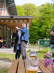 Bild - Eine fröhliche Tanzveranstaltung im Freien, bei der mehrere Paare auf einer Wiese tanzen. Im Hintergrund spielt eine Sängerin mit Gitarre. Tische mit Getränken und Blumen dekorieren den vorderen Bereich. Umgeben von grüner Natur und einem historischen Gebäude.