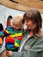 Bild - Eine Frau hält ein kleines Kind, das an einer interaktiven Wand mit Löchern spielt. Das Kind trägt eine bunte Regenjacke. Beide scheinen neugierig und erfreut zu sein, während sie die Ausstellung erkunden.