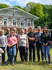 Bild - Eine Gruppe von 14 Personen steht lächelnd auf einer Wiese vor einem historischen Gebäude mit Holzverzierungen. Im Hintergrund sind Bäume und blauer Himmel sichtbar. Die Menschen tragen legere Kleidung und haben unterschiedliche Altersgruppen und Geschlechter.
