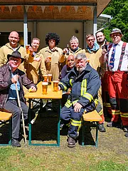 Bild - Eine Gruppe von zwölf Männern steht und sitzt an einem Tisch im Freien, alle tragen handwerkliche Kleidung. Einige halten Bier in den Händen und lachen. Die Szene wirkt fröhlich und gesellig, umgeben von einer grünen Wiese und einem sonnigen Himmel.