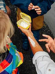 Bild - Eine Person zeigt einer Gruppe von Kindern eine gelbe Schachtel mit einem weißen Pulver, während sie das Pulver auf den eigenen Arm streicht. Die Kinder schauen interessiert zu, wobei eines ein buntes Regenjacke trägt.