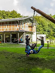 Bild - Drei Kinder spielen auf einer großen Schaukel im Freien, umgeben von einer grünen Wiese und Bäumen. Im Hintergrund ist ein modernes, mehrstöckiges Gebäude zu sehen, in dem sich mehrere Personen aufhalten. Die Szene vermittelt eine fröhliche, aktive Atmosphäre.