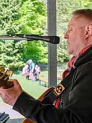 Bild - Ein Mann spielt Gitarre und singt in ein Mikrofon, während er auf einer Bühne steht. Im Hintergrund sind verschwommen Menschen sichtbar, die im Freien bei sonnigem Wetter stehen. Der Mann trägt einen schwarzen Pullover und einen Schal.
