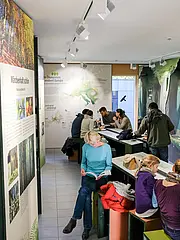 Bild - In einem Raum mit großen Informationswänden sitzen mehrere Menschen an Tischen. Einige lesen, andere unterhalten sich. Eine Frau in einem blauen Oberteil schaut in ein Buch. Die Atmosphäre ist entspannend und lehrreich, mit Fokus auf Bäume und Natur.