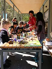 Bild - Eine Gruppe von Kindern, die an einem Tisch im Freien sitzt, bastelt mit verschiedenen Materialien. Eine Betreuerin zeigt ihnen, wie sie kreative Projekte umsetzen können. Der Tisch ist mit verschiedenen Malutensilien und Pflanzen dekoriert, während die Natur im Hintergrund sichtbar ist.
