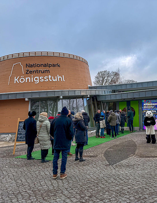 Eine Gruppe von Menschen steht in einer langen Schlange vor dem Eingang des Nationalpark Zentrums Königsstuhl. Das Gebäude hat eine moderne Architektur mit einer markanten runden Fassade. Banner zeigen Informationen über den Park. Der Himmel ist bewölkt.