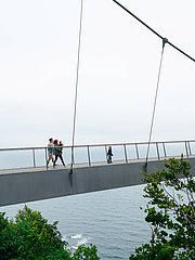 Eine Gruppe von Menschen geht über eine schmale Hängebrücke, die über einen Abgrund und das Meer führt. Links und rechts der Brücke sind Bäume und grünes Gelände sichtbar. Es ist bewölkt und die Szenerie vermittelt eine ruhige, natürliche Atmosphäre.