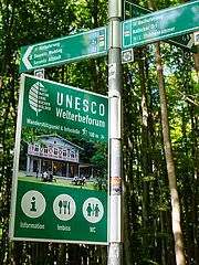 Bild - Ein grünes Schild im Wald, das auf das UNESCO-Welterbe und einen Wanderstützpunkt hinweist. Es zeigt Informationen über einen Imbiss und WC-Einrichtungen. Das Schild zeigt auch eine Abbildung eines Gebäudes, das als Informationsstelle dient.