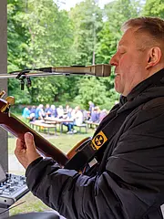 Bild - Ein Musiker steht auf einer Bühne im Freien und spielt Gitarre. Er singt ins Mikrofon und trägt eine dicke Jacke. Im Hintergrund sind einige Menschen zu sehen, die an Tischen sitzen und die Musik genießen. Umgeben von grünen Bäumen schafft die Szene eine gesellige Atmosphäre.