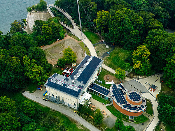 Eine Luftaufnahme zeigt ein modern gestaltetes Gebäude mit Solarzellen, umgeben von Bäumen und Grünflächen. In der Nähe verläuft ein geschwungener Weg, der zu einer Aussichtsplattform an einer Klippe führt, die auf das Meer blickt.