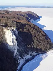 Eine winterliche Landschaft mit einer weißen, gefrorenen Küstenlinie. Ein Steilhang mit weißen Kreidefelsen und kahlen Bäumen ist im Vordergrund sichtbar, während die Weite des gefrorenen Ozeans und der klare Himmel im Hintergrund zu sehen sind.