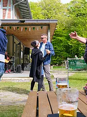 Bild - Eine Gruppe von Menschen tanzt fröhlich im Freien vor einem Gebäude. Im Hintergrund spielt eine Band Musik. Auf einem Tisch im Vordergrund stehen Gläser mit Getränken und eine Vase mit frischen Blumen. Die Atmosphäre ist lebhaft und gesellig.