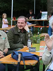 Bild - Eine Gruppe von Männern sitzt an einem Tisch, während ein älterer Mann mit grauem, lockigem Haar applaudiert. Die Umgebung zeigt Tische unter Bäumen, einige Plakate im Hintergrund und eine kleine Pflanze in einem Topf auf dem Tisch.
