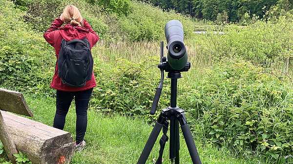 Eine Person mit einem Rucksack steht auf einer Wiese und schaut in die Ferne. Neben ihr steht ein Stativ mit einem Fernrohr, das auf die Landschaft gerichtet ist. Im Hintergrund sind Bäume und eine grüne Umgebung zu sehen.