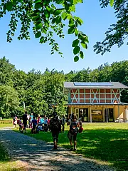Bild - Eine Gruppe von Menschen geht einen Weg entlang zu einem modernen Gebäude mit Holz- und Glasschnitt. Das Gebäude steht in einer grünen Umgebung mit Bäumen und Wiese unter einem blauen Himmel. Es scheint ein Freizeit- oder Bildungsort zu sein.