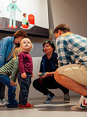 Eine Gruppe von Erwachsenen und Kindern interagiert in einem modernen Raum. Zwei Kinder stehen im Mittelpunkt, während ein Erwachsener sich zu ihnen niederbeugt und mit ihnen spricht. Im Hintergrund ist eine grafische Wandgestaltung sichtbar.