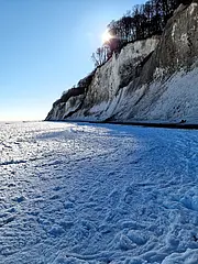 Bild - Ein schneebedeckter Strand mit hohen, weißen Klippen im Hintergrund. Die Sonne scheint über die Szene, und der Himmel ist klar und blau. Der Schnee bedeckt den Boden und es gibt deutlich sichtbare Spuren im Schnee.