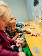 Bild - Zwei Kinder stehen an einem Tisch mit Ausstellungsobjekten. Das Mädchen mit blonden Haaren beugt sich vor, während der Junge mit kurzen Haaren interessiert schaut. In der Nähe sind Behälter mit kleinen Tieren, die sie aufmerksam betrachten.