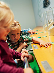 Zwei Kinder stehen an einem Tisch mit Ausstellungsobjekten. Das Mädchen mit blonden Haaren beugt sich vor, während der Junge mit kurzen Haaren interessiert schaut. In der Nähe sind Behälter mit kleinen Tieren, die sie aufmerksam betrachten.