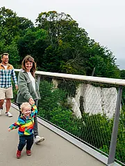 Bild - Eine Familie mit zwei kleinen Kindern spaziert auf einem Weg mit Geländer. Die Erwachsenen, ein Mann und eine Frau, gehen neben den Kindern, die fröhlich sind. Im Hintergrund sind Bäume und das Meer zu sehen, unter einem grauen Himmel.