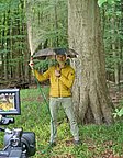 Ein Mann steht unter einem Baum im Wald und hält einen Regenschirm. Er hat einen grünen Schlauch in der Hand, aus dem Wasser spritzt. Er trägt eine gelbe Jacke und graue Hosen. Im Vordergrund steht eine Kamera, die ihn filmt.
