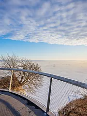 Bild - Ausblick von einem Aussichtspunkt auf eine winterliche Landschaft mit gefrorenem Wasser und einem klaren Himmel. Die Sonne scheint am Horizont, während sich eine sanfte Kurve der Umzäunung um den Punkt zieht.
