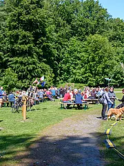 Bild - Eine Veranstaltung im Freien mit vielen Menschen, die an Tischen sitzen und miteinander sprechen. Im Vordergrund spielen Kinder an Spielgeräten, während Erwachsene sich unterhalten. Eine Hund läuft durch die Szene. Im Hintergrund sind Bäume und eine informative Wandtafel sichtbar.