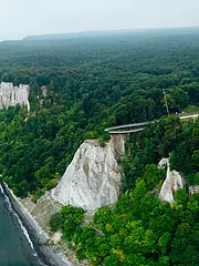 Bild - Luftaufnahme einer Küstenlandschaft mit steilen, weißen Klippen und dichten Wäldern. Im Hintergrund ist ein Teil der Landschaft mit einer kleinen Struktur zu sehen, die sich nahe der Klippe befindet. Das Meer erstreckt sich entlang der Küste.