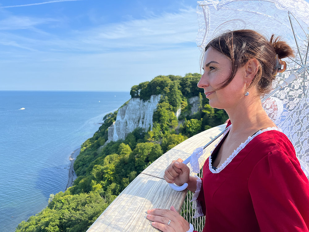 Eine Frau in einem rot-weißen Kleid steht auf einer Aussichtsplattform und schaut auf das Meer. Sie hält einen Regenschirm in der Hand und der Blick ist auf die Küste mit bewaldeten Klippen gerichtet. Der Himmel ist klar und blau.